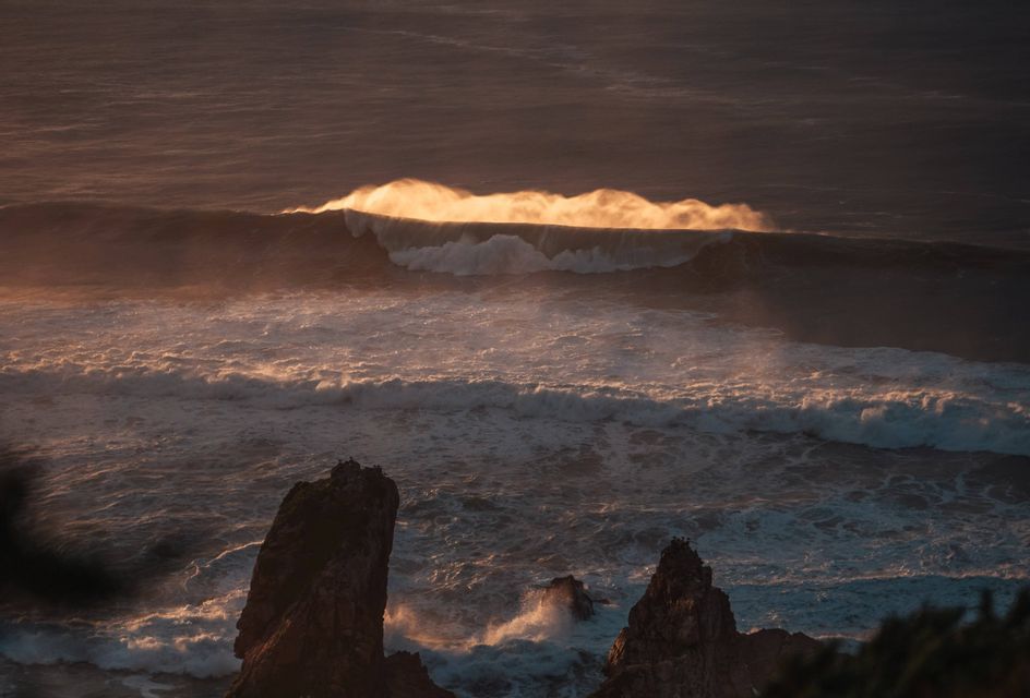 Una grande onda oceanica si infrange nell'acqua, la sua cresta che risplende di luce solare dorata sopra le rocce scure in primo piano.