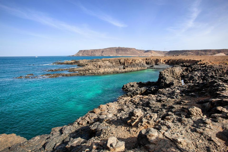 Une côte déchiquetée et rocheuse encadre une étroite crique aux eaux turquoise éclatantes, sous un ciel bleu parsemé de légers nuages.