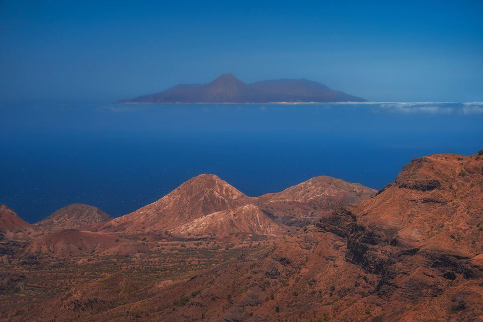 Vue depuis des collines volcaniques rouges sur une mer d'un bleu profond vers une île lointaine flottant sur une basse bande de nuages.