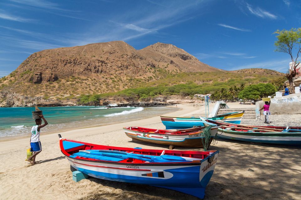 Une personne porte un panier sur sa tête, marchant le long de bateaux de pêche colorés sur une plage de sable avec une grande montagne en arrière-plan.