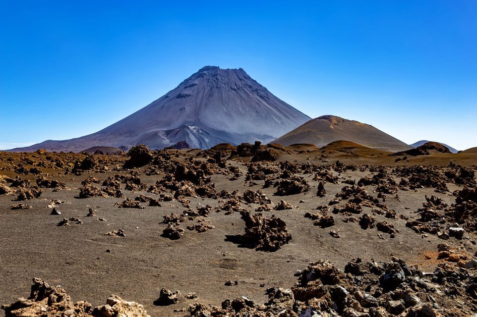 Un grand volcan se dresse d'un paysage aride de sable sombre et de roches de lave rougeâtres sous un ciel bleu clair.