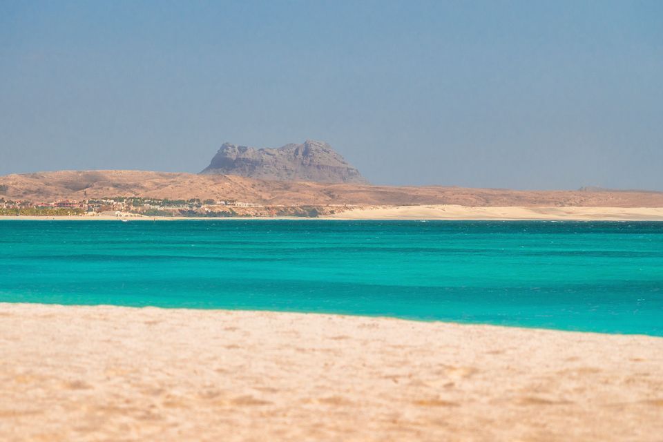Vue d'une plage de sable fin à travers des eaux turquoise vers une côte aride lointaine avec une grande formation rocheuse sous un ciel clair.