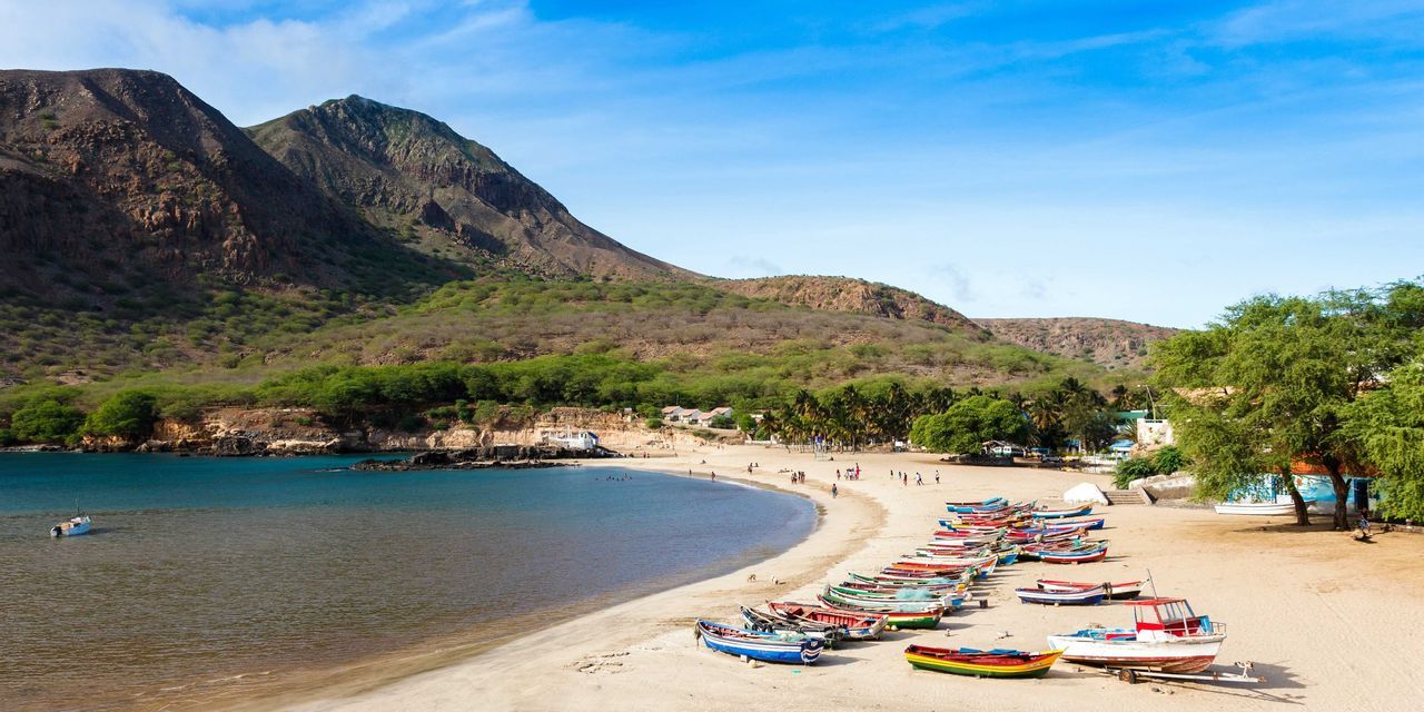 Une flotte de bateaux de pêche colorés repose sur une plage de sable dans une baie, avec des montagnes vertes en arrière-plan.