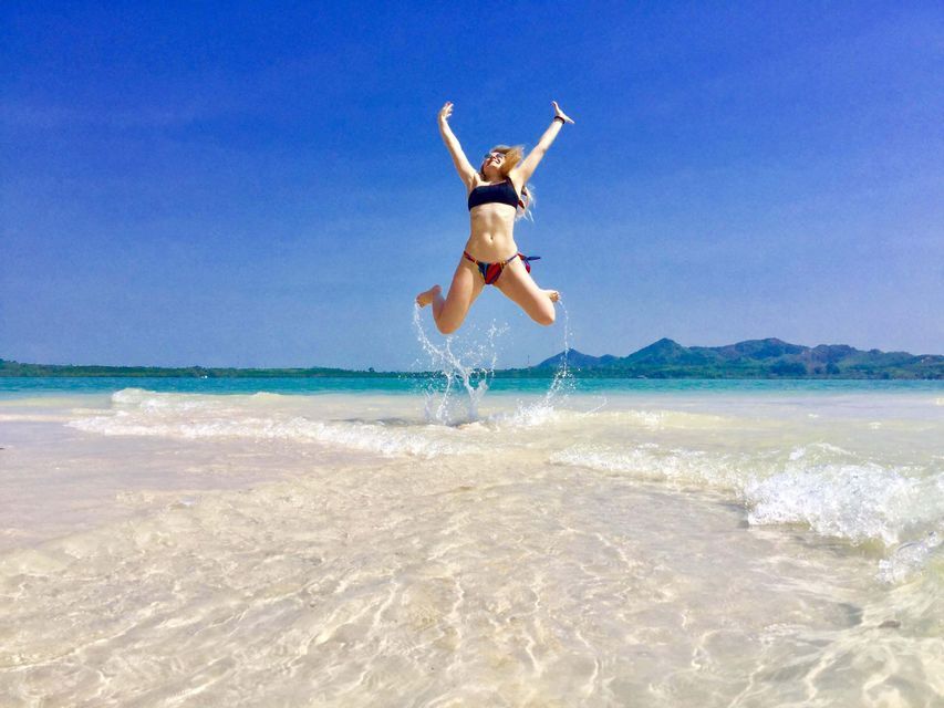 A woman in a bikini jumps out of the shallow, clear water at a tropical beach, with her arms raised towards the blue sky.