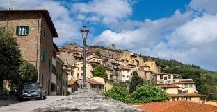 Un borgo collinare con tradizionali edifici in pietra e un lampione, visto da una strada selciata sotto un cielo azzurro con nuvole.