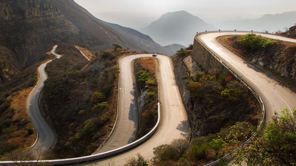Un'unica auto percorre una strada tortuosa, con tornanti a gomito, attraverso un paesaggio montuoso ripido e avvolto nella foschia.