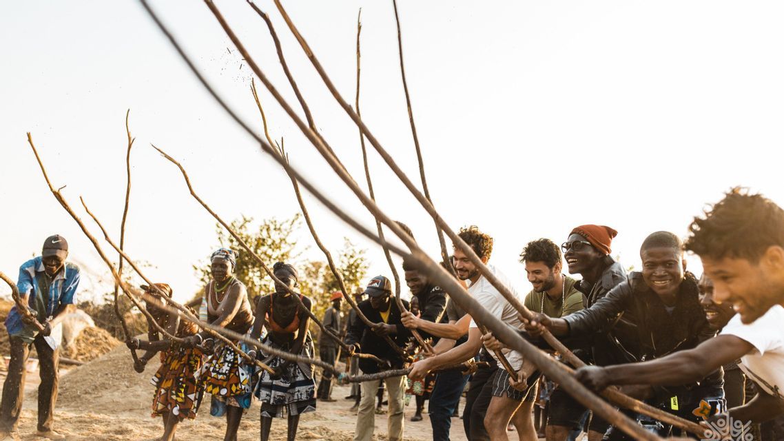Un viaggio di gruppo WeRoad e persone del posto sorridono e ridono insieme mentre partecipano a un'attività tradizionale con lunghi bastoni all'aperto.
