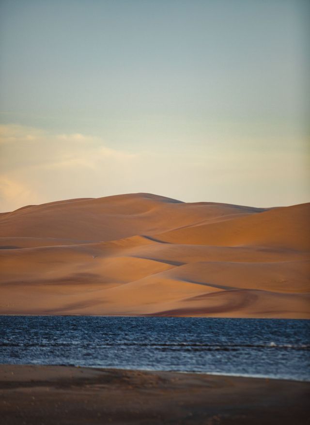 Dune di sabbia dorata si ergono dal bordo di uno specchio d'acqua blu scuro sotto un cielo pallido.