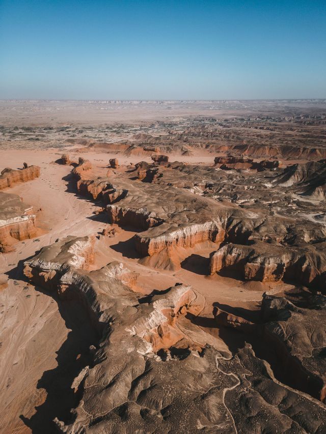 Vista aerea di un vasto paesaggio desertico con canyon marrone-rossastro e formazioni rocciose stratificate sotto un cielo limpido.