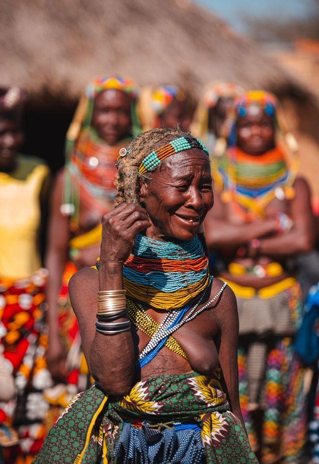 Una donna anziana sorridente indossa un abito tradizionale colorato, inclusi collane, braccialetti e una fascia per capelli intricati.