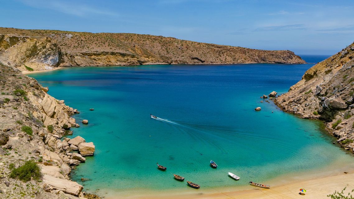 Una vista aerea di un motoscafo che lascia una scia in una baia turchese circondata da scogliere aride e rocciose e una piccola spiaggia sabbiosa.