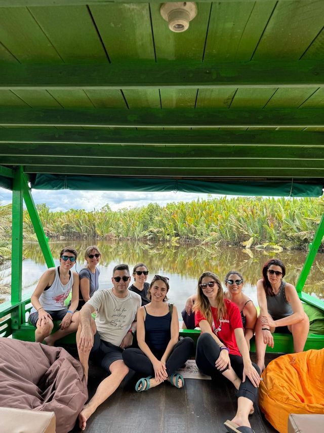 Un grupo de viaje de WeRoad está en la cubierta de un barco de madera verde, posando para una foto en un río con vegetación exuberante.