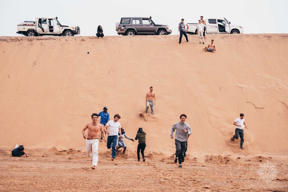 Uomini di un viaggio di gruppo WeRoad che corrono e scivolano giù da una grande duna di sabbia, con veicoli fuoristrada parcheggiati sulla cresta.