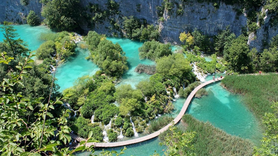 Ein erhöhter Blick auf einen Holzsteg, der sich über türkisfarbene Seen und Wasserfälle schlängelt, umgeben von üppiger Vegetation und einer Felswand.