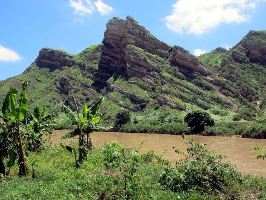 Lussureggianti montagne verdi con formazioni rocciose stratificate si innalzano su un fiume fangoso, con alberi di banane sulla sponda sotto un cielo azzurro.