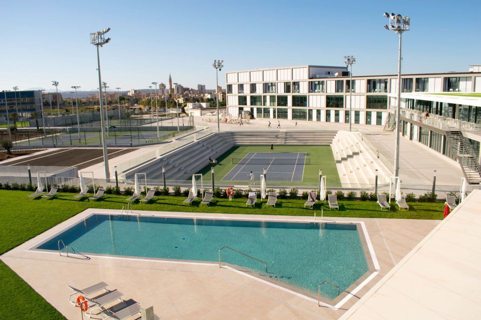 Veduta dall'alto di un moderno complesso sportivo con una piscina in primo piano e un campo da tennis con giocatori sullo sfondo.