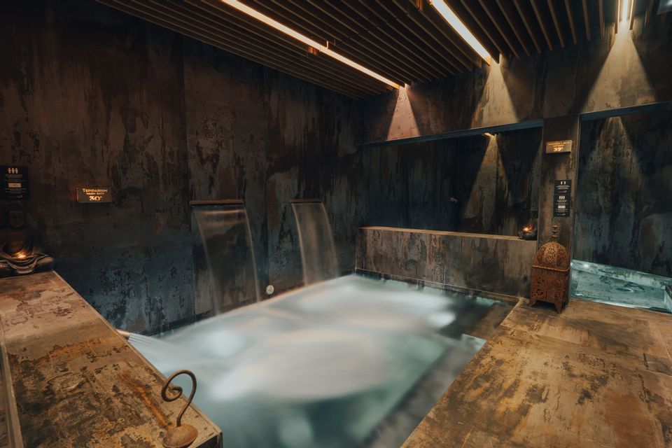 Piscina termale interna con piccole cascate, circondata da pareti scure e materiche e illuminata da calde luci a soffitto.