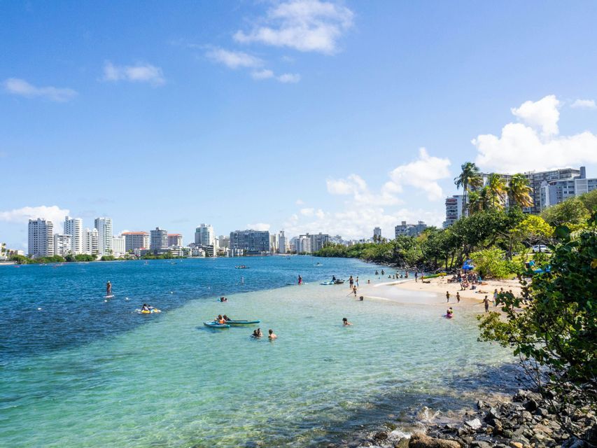 Le persone nuotano e si rilassano nelle limpide acque turchesi di una baia, con una spiaggia sabbiosa e lo skyline di una città sullo sfondo.