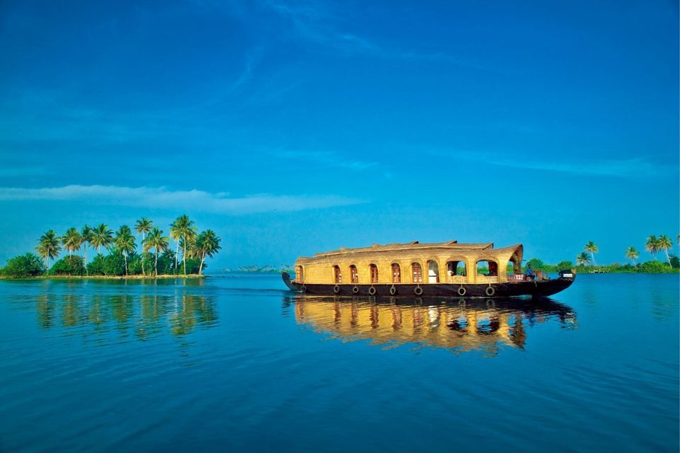 Ein traditionelles Hausboot schwimmt auf ruhigem, blauem Wasser, das sich auf der Oberfläche neben einer mit Palmen gesäumten Insel spiegelt.