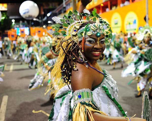 Une femme vêtue d'un costume de carnaval vert et blanc éclatant avec une grande coiffe sourit à l'appareil photo lors d'une parade nocturne.