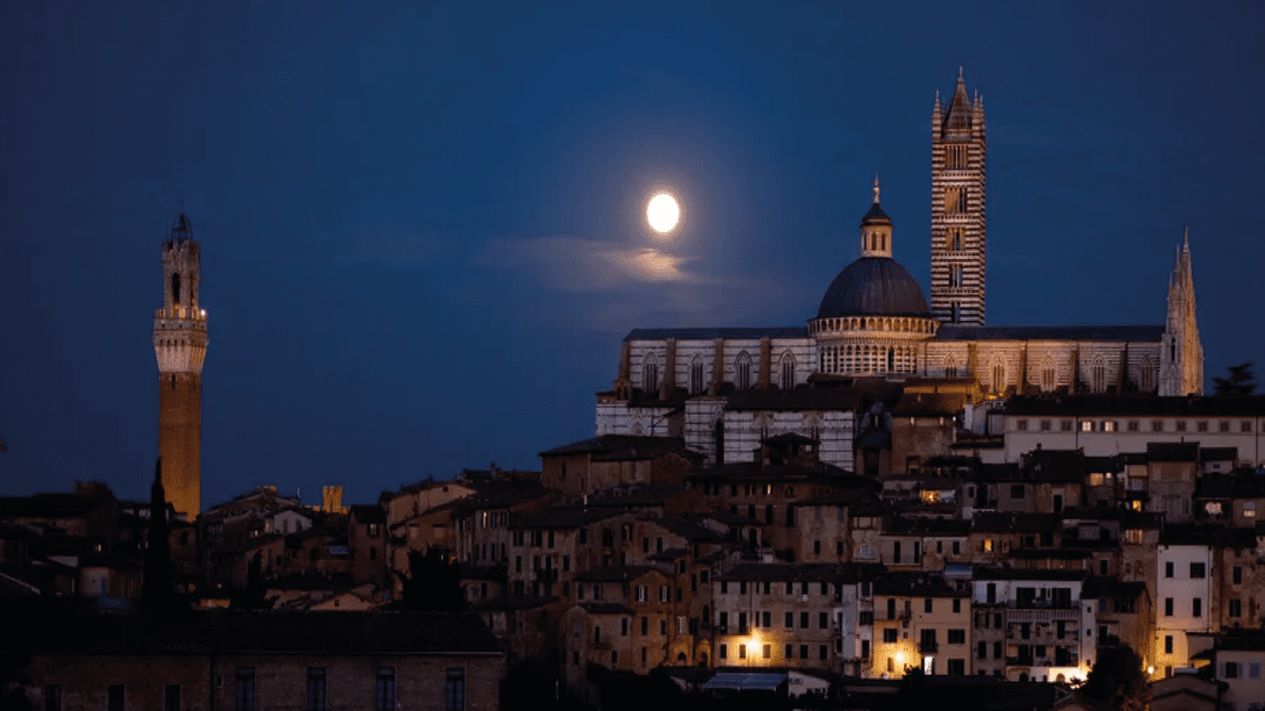 Una città storica con una cattedrale e torri illuminate, sotto un cielo notturno buio con una luna piena splendente.