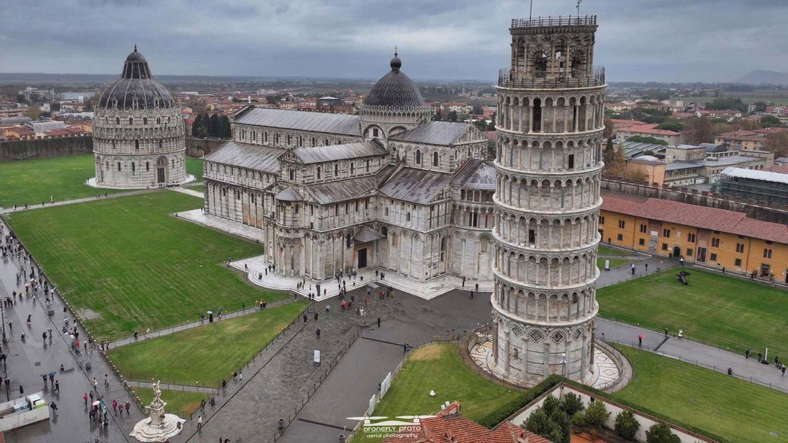 Una vista aerea di una piazza storica con una torre pendente, una cattedrale e un battistero circondati da prati verdi in una giornata nuvolosa.