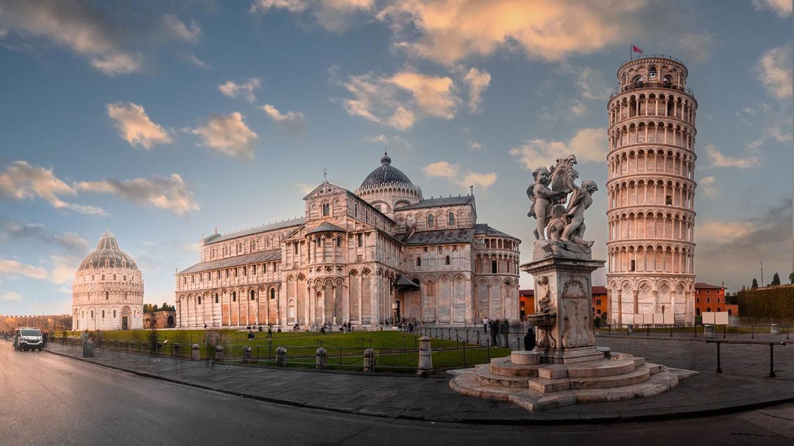 Una vista panoramica di una grande cattedrale, una torre pendente e un battistero accanto a un prato verde sotto un cielo parzialmente nuvoloso.