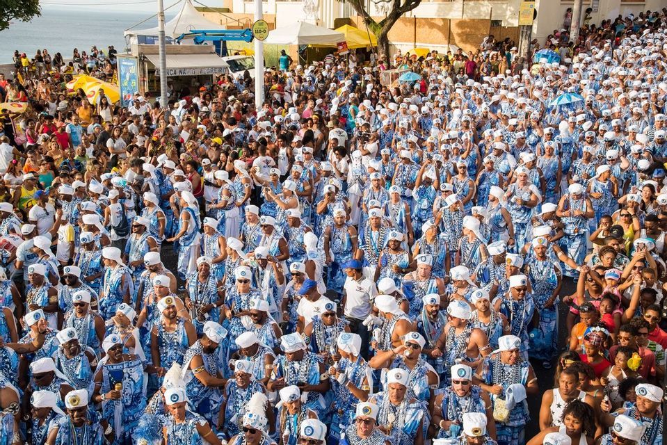 Un grand groupe de personnes en costumes assortis bleus et blancs défile dans une rue bondée près de la mer.