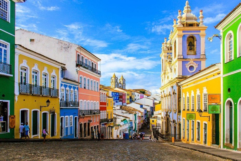 Une rue pavée ensoleillée est bordée de bâtiments coloniaux aux couleurs vives et d'une grande église sous un ciel bleu.