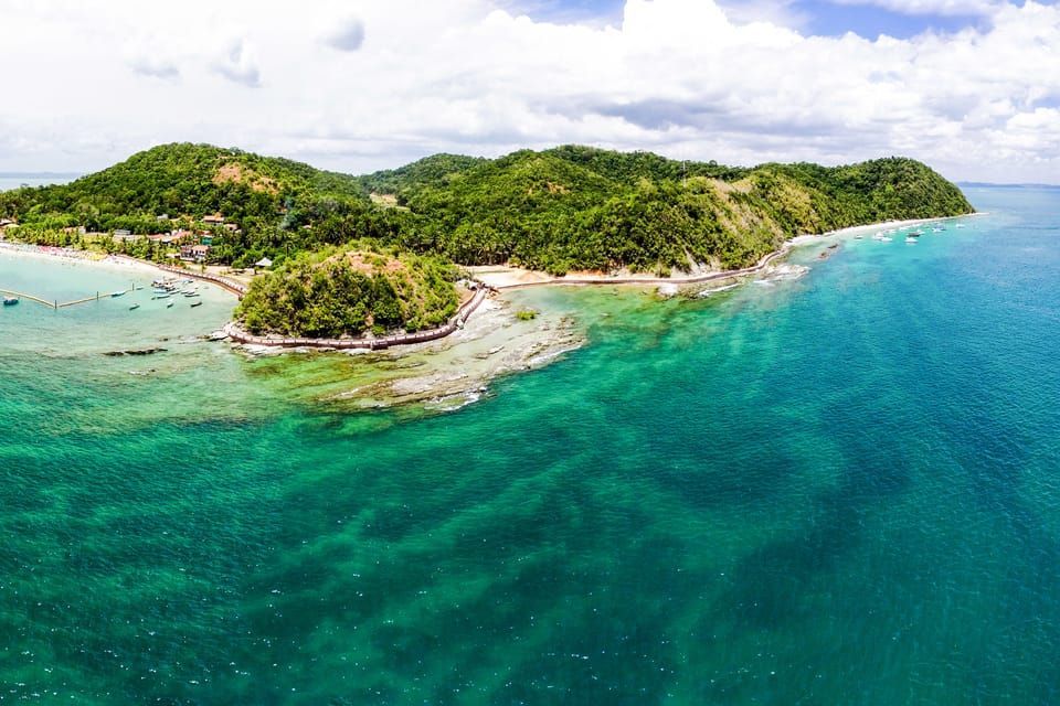 Vue aérienne d'une île tropicale avec des collines verdoyantes, une eau turquoise claire et une petite baie avec des bateaux.