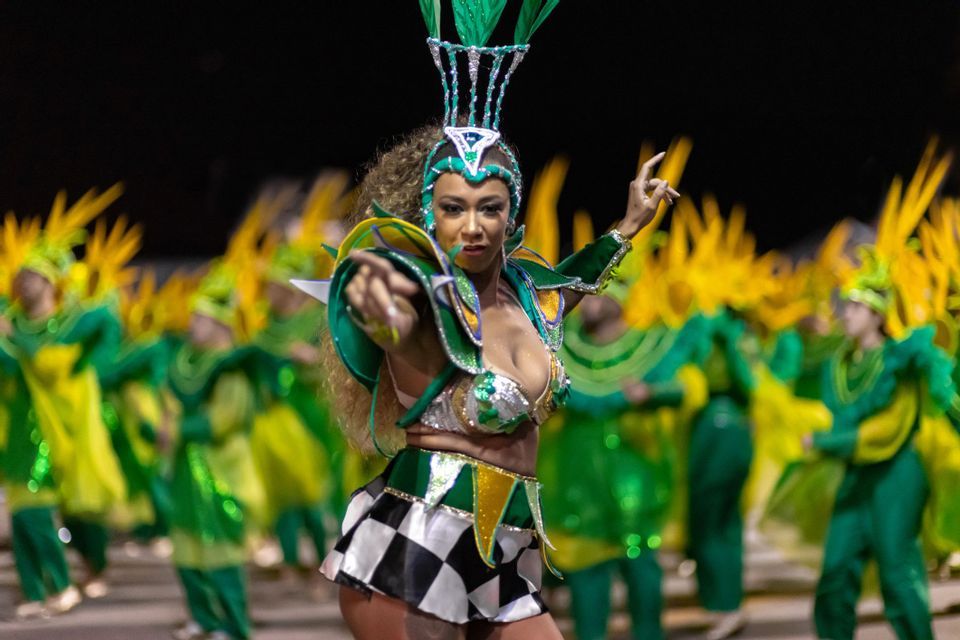 Une femme en costume de carnaval vert et argenté orné de plumes danse et pointe vers l'avant lors d'une parade nocturne.