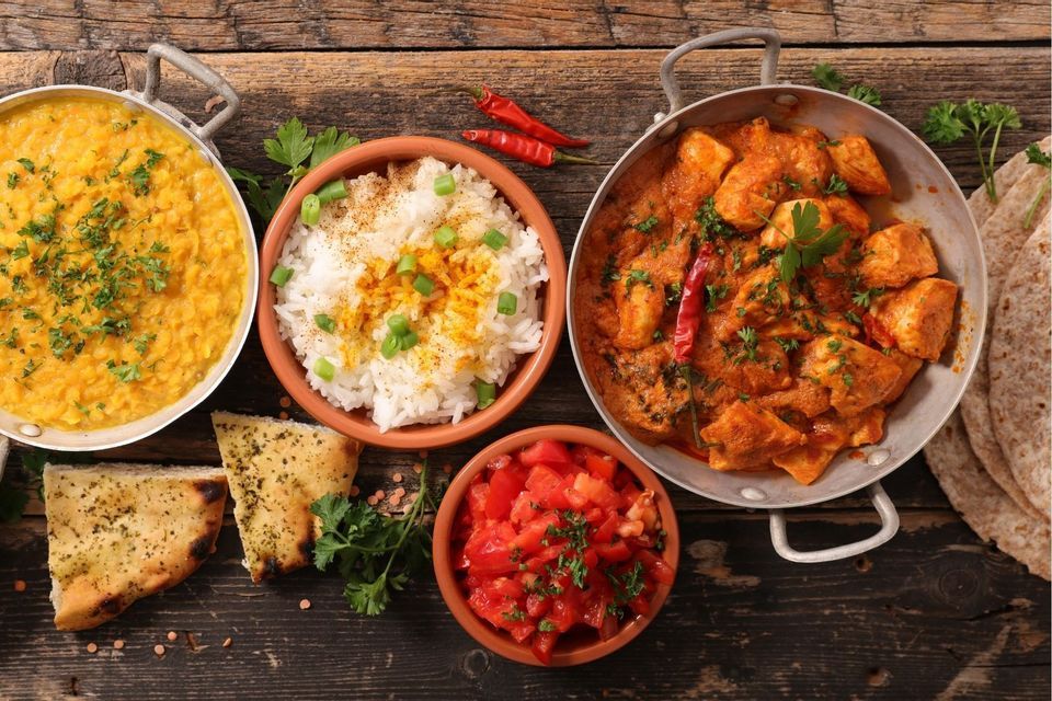 Eine Vogelperspektive auf indische Gerichte, darunter Hühnercurry, Dal, Reis, Salat und Brot, angerichtet auf einem dunklen Holztisch.