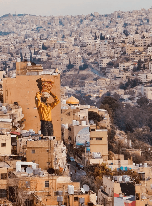 Un grande murale raffigurante un uomo con una camicia gialla che trasporta un capitello sulla testa, dipinto su un edificio che domina una città collinare densamente popolata.