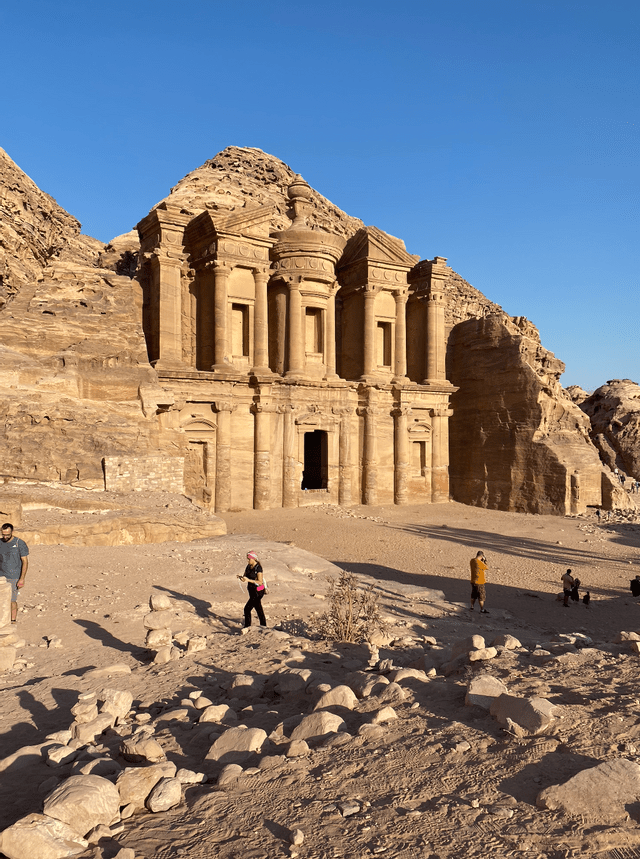 Un tempio monumentale scavato nella scogliera di arenaria, con persone in primo piano tra le rocce sotto un cielo azzurro e limpido.