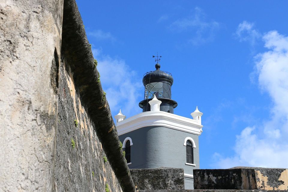 Una torre faro grigia con la cima nera vista da dietro un vecchio muro di pietra sotto un cielo blu.