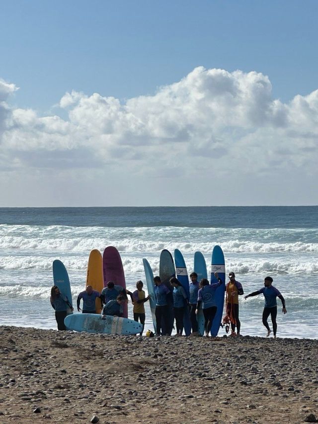 Un viaggio di gruppo WeRoad in posa con tavole da surf colorate su una spiaggia di ciottoli con l'oceano sullo sfondo.