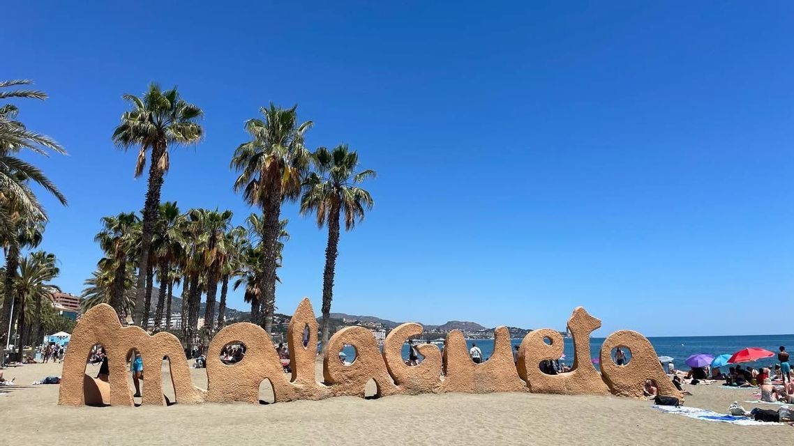 Une sculpture couleur sable formant « Malagueta » se dresse sur une plage ensoleillée, avec des palmiers et un ciel bleu clair.
