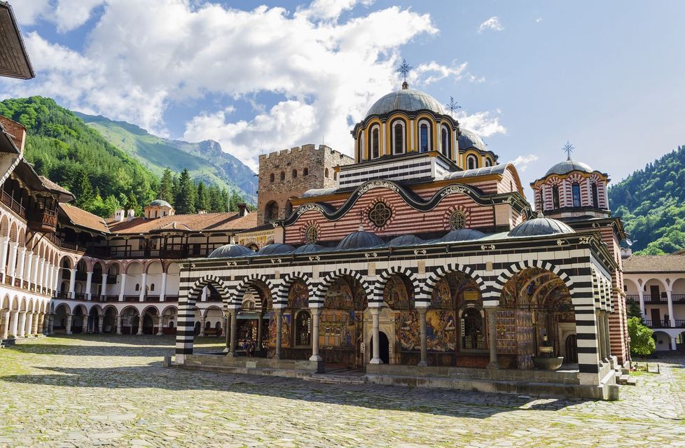 Un monastero ornato con archi e cupole a strisce in un cortile acciottolato, con montagne boscose sotto un cielo nuvoloso.