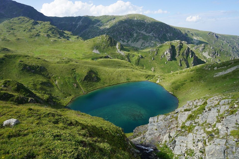 Un lago alpino di un blu limpido si trova in una conca circondata da dolci montagne verdi sotto un cielo con nuvole leggere.