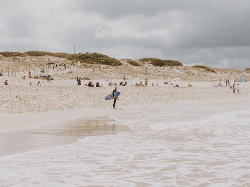 Un surfista con traje de neopreno sostiene una tabla de surf azul a la orilla del agua en una playa concurrida con dunas de arena bajo un cielo nublado.