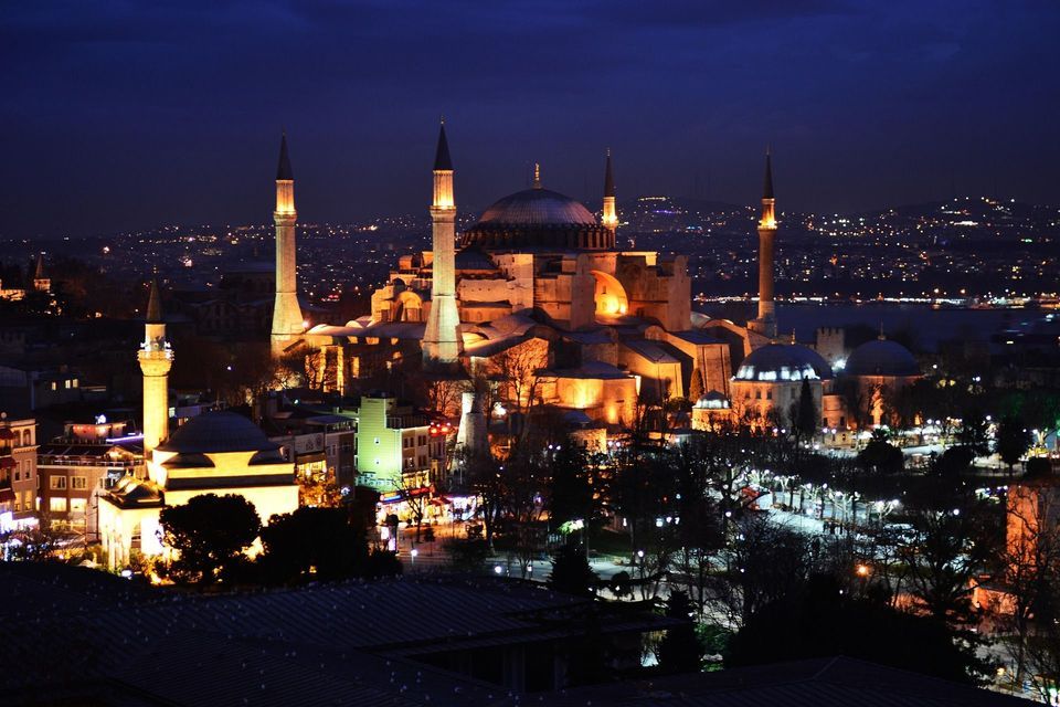 Una grande moschea con cupola e minareti illuminati da luci dorate di notte, sullo sfondo di un vasto skyline cittadino.