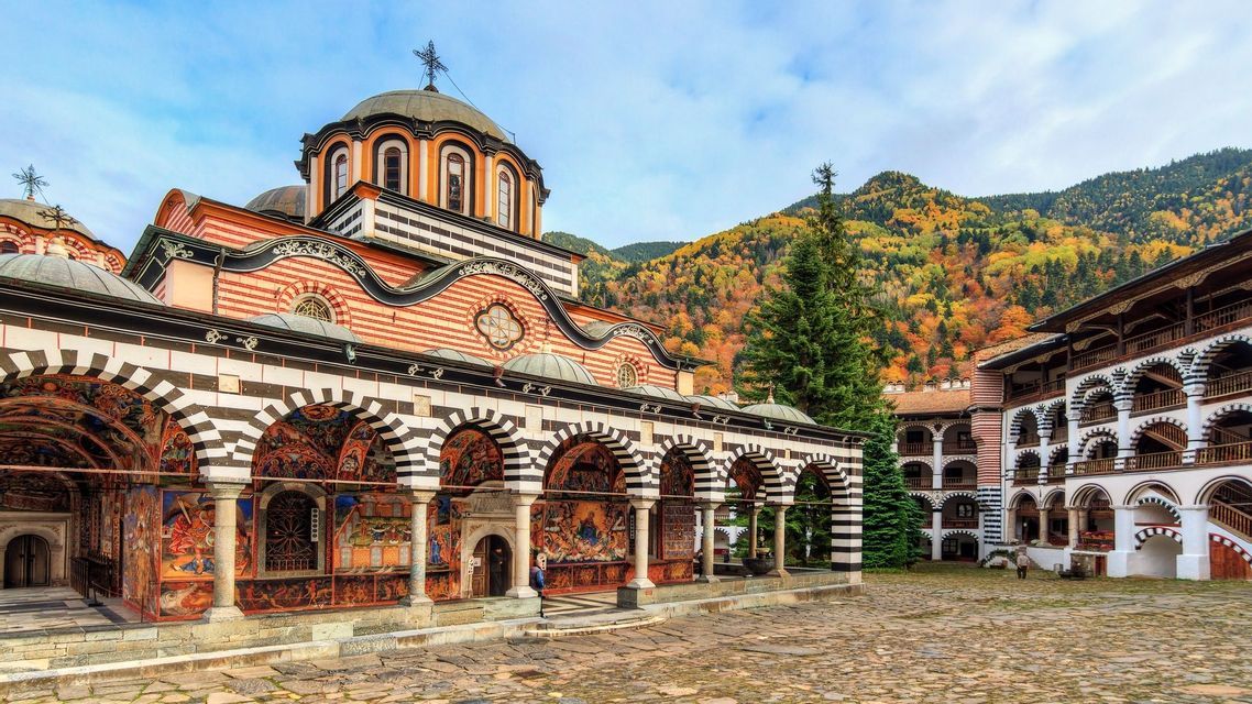 Un sontuoso monastero con archi a strisce e affreschi dettagliati in un cortile, e una montagna coperta di alberi autunnali sullo sfondo.