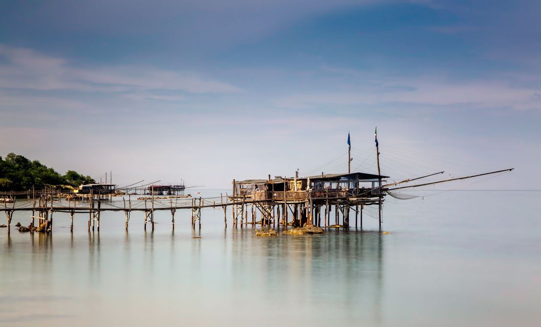 Piattaforme da pesca in legno su palafitte, con grandi reti, si ergono su acque calme e riflettenti vicino a una riva alberata.