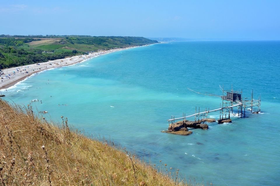 Vista dall'alto di una costa con una lunga spiaggia, acqua turchese e un tradizionale molo da pesca in legno su palafitte.