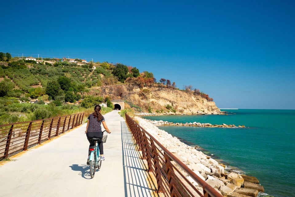 Una donna in bicicletta su un sentiero pavimentato lungo una costa rocciosa con acqua turchese, in direzione di un tunnel.