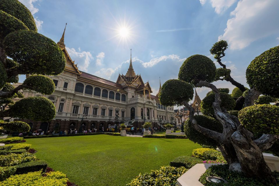 Un palais orné au toit doré se dresse derrière une vaste pelouse impeccablement entretenue, encadré par des arbres taillés en topiaire sous un soleil éclatant.