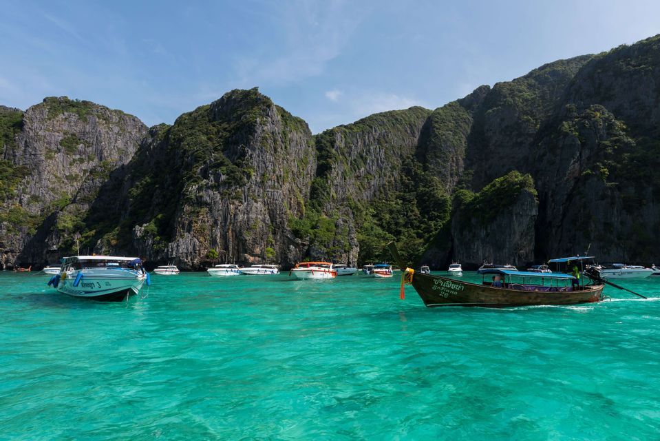 Plusieurs bateaux touristiques, dont une pirogue à longue queue, sur des eaux turquoise dans une baie entourée de falaises rocheuses abruptes aux cimes verdoyantes.