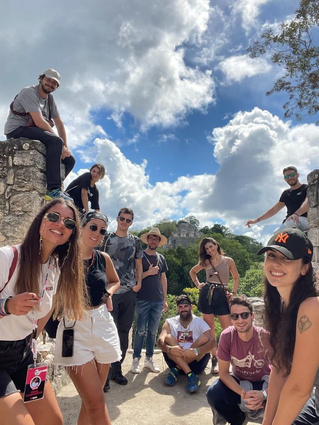 Eine WeRoad-Gruppenreise posiert für ein Foto auf Steinruinen, mit einem Tempel und Dschungel im Hintergrund unter bewölktem Himmel.