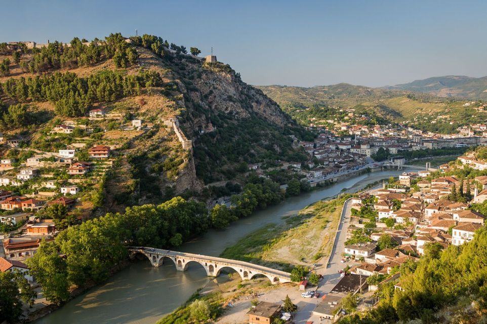 Un pont de pierre voûté enjambant une rivière dans une ville historique nichée dans une vallée, dominée par une colline fortifiée.