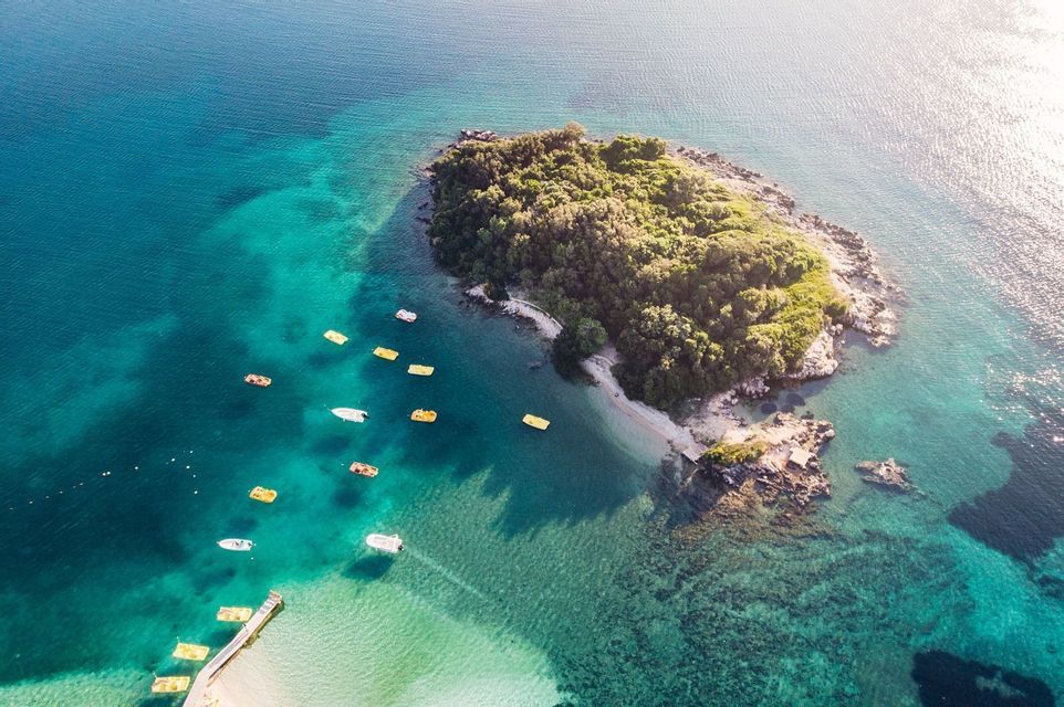 An aerial view of a small, tree-covered island in turquoise water, with several small boats floating near its sandy shore.